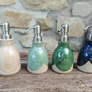 Peut inclure: Quatre distributeurs de savon en céramique avec des pompes en acier inoxydable. Chaque bouteille présente une base arrondie avec une finition crème mouchetée et un vernis coloré distinct sur la partie supérieure, allant du rose clair au vert, vert foncé et bleu.