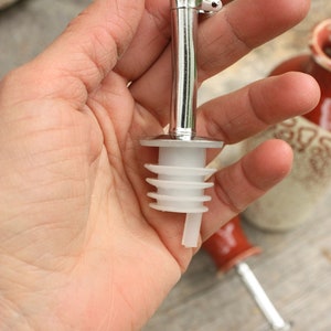 May include: A silver bottle pourer with a white plastic spout. The pourer has a lever to control the flow of liquid.