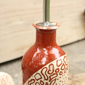 May include: A red and white ceramic oil and vinegar bottle with a stainless steel pour spout. The bottle has a speckled glaze and a geometric pattern.