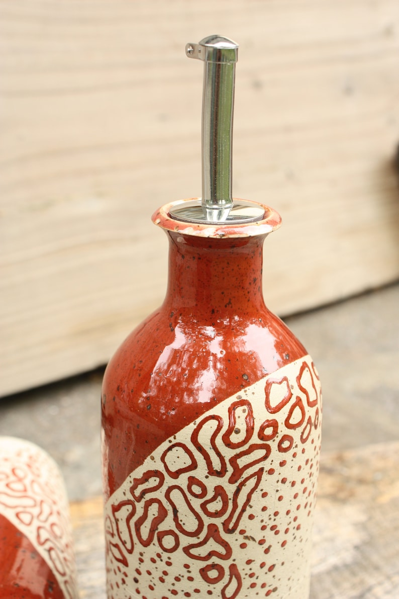 May include: A red and white ceramic oil and vinegar bottle with a stainless steel pour spout. The bottle has a speckled glaze and a geometric pattern.