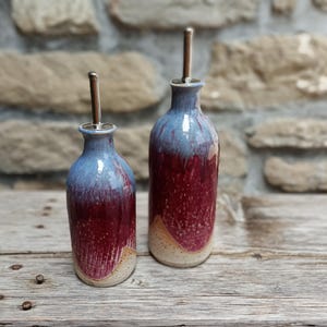 May include: Two ceramic oil dispensers with a gradient glaze featuring a light blue top, a deep magenta middle, and a cream-coloured base. Each bottle has a narrow neck and a stainless steel pour spout inserted into the top opening.
