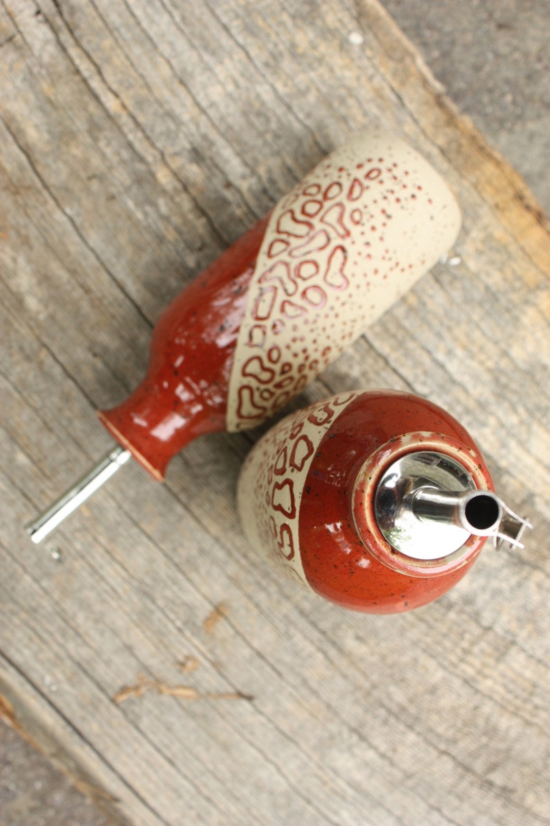 May include: Two ceramic oil and vinegar bottles with red and white speckled glaze and a geometric pattern. The bottles have metal pour spouts.