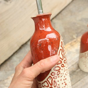 May include: A red and white ceramic oil and vinegar bottle with a silver pour spout. The bottle has a speckled glaze and a white design with red accents.