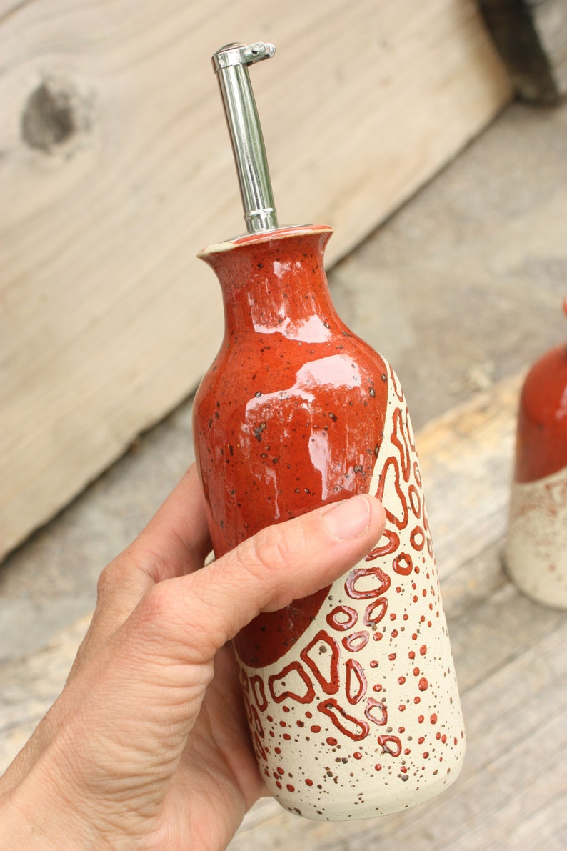 May include: A red and white ceramic oil and vinegar bottle with a silver pour spout. The bottle has a speckled glaze and a white design with red accents.