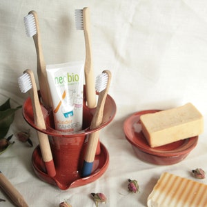 Könnte beinhalten: Ein roter Keramik-Zahnbürstenhalter mit vier Holz-Zahnbürsten und einer Tube Zahnpasta mit der Aufschrift "neobio Kinder Zahngel".
