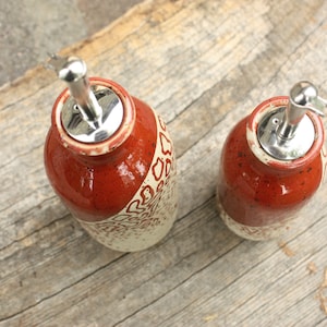 May include: Two ceramic oil and vinegar bottles with red and white speckled glaze and silver pour spouts. The bottles are sitting on a wooden surface.