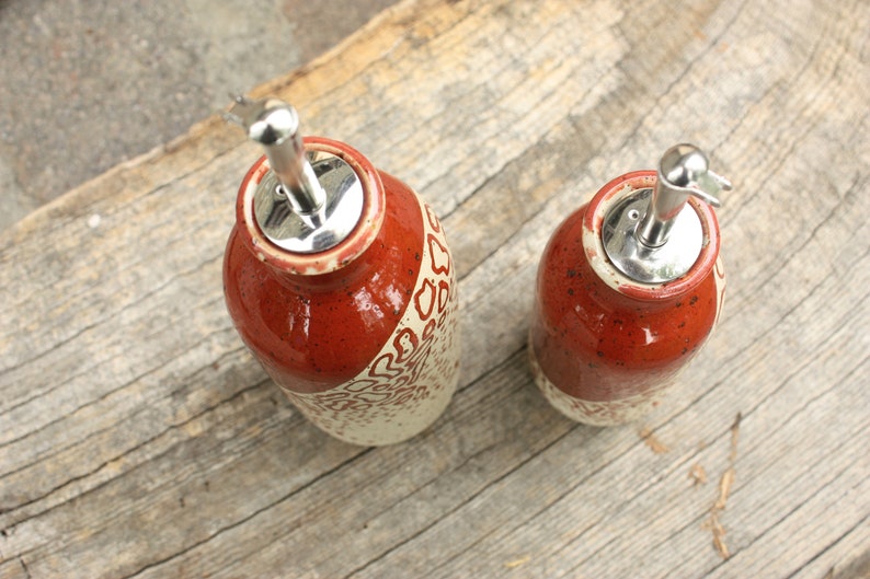 May include: Two ceramic oil and vinegar bottles with red and white speckled glaze and silver pour spouts. The bottles are sitting on a wooden surface.