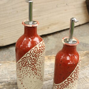 May include: Two ceramic oil and vinegar bottles with stainless steel pour spouts. The bottles are decorated with a white and red speckled glaze and a red and white abstract design.