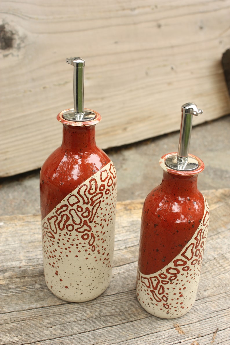 May include: Two ceramic oil and vinegar bottles with stainless steel pour spouts. The bottles are decorated with a white and red speckled glaze and a red and white abstract design.