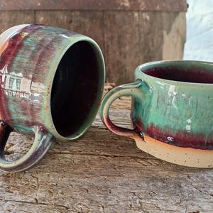 Pode incluir: Duas canecas de cerâmica feitas à mão com uma vidragem única. As canecas apresentam um esquema de cores verde-azulado e bordô com uma base natural sem vidrado. As alças são arredondadas e o interior é de cor bordô profundo. As canecas estão numa superfície de madeira.