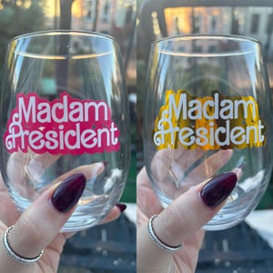 May include: Two clear glass wine glasses with the text "Madam President" in white letters on a pink and gold background.