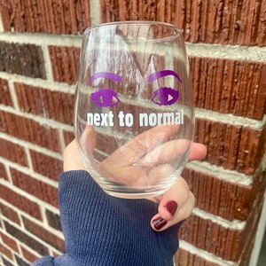 May include: A clear glass wine glass with a purple eye design and the text "next to normal" printed on the front.