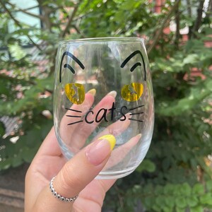 May include: A clear glass wine glass with a black cat face design. The cat has yellow eyes and the word "cats" is written below the face.