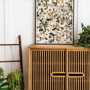 May include: A wooden cabinet with two doors and a black framed print of an abstract floral design hanging above it. The cabinet has a slatted design and is made of light brown wood. The print is a watercolor style with a white background and brown, green, and yellow flowers.