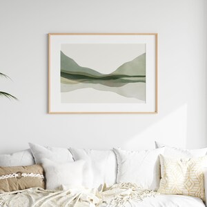 May include: A framed landscape art print featuring a watercolor depiction of rolling green hills and their reflection in a body of water. The artwork is in a light wood frame and hangs above a white sofa with decorative pillows.