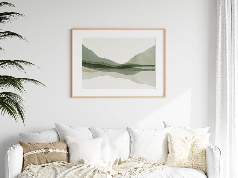 May include: A framed landscape art print featuring a watercolor depiction of rolling green hills and their reflection in a body of water. The artwork is in a light wood frame and hangs above a white sofa with decorative pillows.