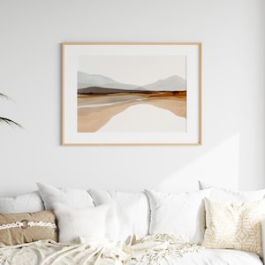 May include: A framed abstract landscape print with a light brown wood frame. The print features a watercolor style depiction of a mountain range with a light blue sky and brown and beige hills.