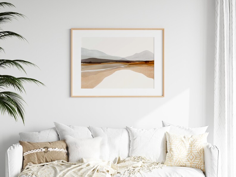 May include: A framed abstract landscape print with a light brown wood frame. The print features a watercolor style depiction of a mountain range with a light blue sky and brown and beige hills.