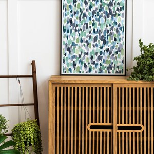 Puede incluir: Un armario de madera con dos puertas y una impresi&oacute;n enmarcada en negro de una pintura abstracta de acuarela con colores azules y verdes. El armario tiene un dise&ntilde;o de listones y est&aacute; sentado sobre una superficie blanca.