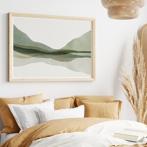 May include: A framed watercolor print featuring an abstract landscape in shades of green and gray. The artwork is mounted in a light wood frame and hangs above a bed with mustard-colored bedding and white pillows. A decorative lamp and dried pampas grass are also visible.