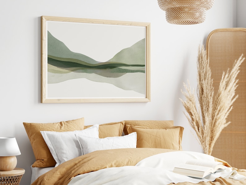 May include: A framed watercolor print featuring an abstract landscape in shades of green and gray. The artwork is mounted in a light wood frame and hangs above a bed with mustard-colored bedding and white pillows. A decorative lamp and dried pampas grass are also visible.