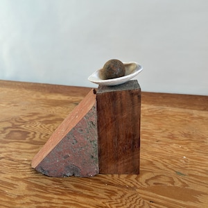 Puede incluir: Una composición de naturaleza muerta con una pequeña esfera texturizada en un cuenco blanco en forma de concha. El cuenco está colocado sobre un bloque de madera oscura, con un trozo de piedra triangular de color marrón rojizo a la izquierda. El fondo es liso y claro.