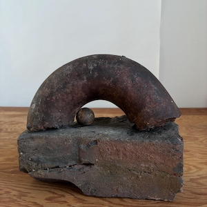 Può includere: Scultura astratta rustica con un elemento metallico curvo a forma di arco su una base di mattoni rettangolare. Una piccola sfera è posta sotto l'arco. La scultura ha un aspetto invecchiato con toni rosso-marrone e grigi.