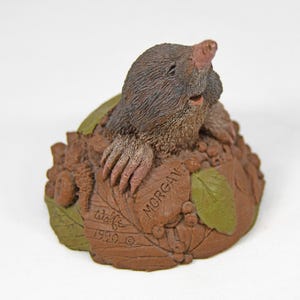 May include: A detailed figurine of a mole emerging from a mound of earth, with a pink nose and detailed fur. The base includes a leaf with the name "Morgan" and the year "1990" inscribed. The figurine is brown and green.