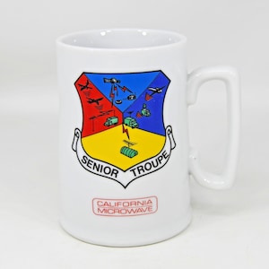 May include: White ceramic mug featuring a colorful emblem with the words "SENIOR TROUPE" and "CALIFORNIA MICROWAVE." The emblem has red, blue, and yellow sections with various illustrations. The mug has a large, square handle.