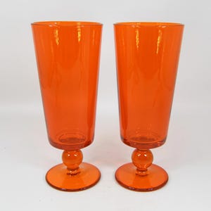 May include: Two tall, orange-colored drinking glasses. Each glass has a flared top, a round base, and a spherical detail on the stem. The glasses are made of transparent glass and are set against a plain background.