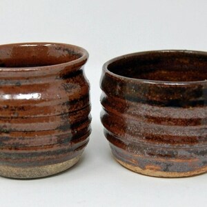 May include: Two ceramic cups with a glossy, dark brown glaze and horizontal ribbed textures. The cups feature a rounded shape with a slightly flared rim and a visible, unglazed tan base at the bottom of each vessel, showcasing a handcrafted appearance.