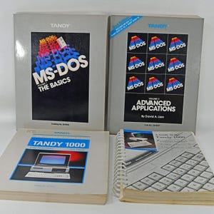 Op de afbeelding: Vier vintage Tandy computerhandleidingen waaronder MS-DOS The Basics, MS-DOS Advanced Applications, The Complete Guide to the Tandy 1000 en een spiraalgebonden Tandy 1000 gids. De boeken hebben grijze covers, blauwe accenten en technische tekst.