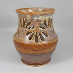 May include: A handcrafted ceramic vase with a unique design. The vase features a warm color palette of brown, orange, and tan hues. Decorative cut-out floral patterns adorn the upper portion of the vase, adding visual interest to this decorative piece.