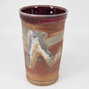 Puede incluir: Un vaso de cerámica hecho a mano con una superficie texturizada. La taza presenta un borde burdeos oscuro, con bandas de color beige, gris y blanco, y un diseño abstracto central en azul y blanco. La base es de color beige claro.