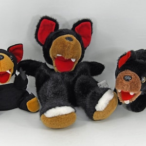 May include: Three plush Tasmanian devil toys. The toys are black with tan and red accents. The toys have open mouths with white teeth and red tongues. The toys are in various poses, with one lying on its back.