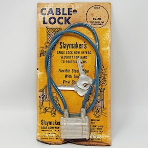 May include: A vintage cable lock on its original packaging. The packaging is yellowed with age and features the text "CABLE LOCK" and "Slaymaker's". The lock itself is silver and blue, with a key inserted. The packaging also includes illustrations.