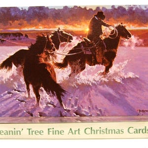 Op de afbeelding: Een kerstkaart met een schilderij van een cowboy te paard die een stier vangt in een besneeuwd landschap bij zonsondergang. De kaart wordt weergegeven in een transparante plastic standaard. De tekst "Leanin' Tree Fine Art Christmas Cards" is zichtbaar.