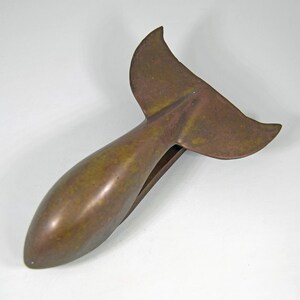 May include: A bronze-colored whale tail paper clip. The clip has a rounded body and a tail fin design. The metal has a slightly aged appearance, with variations in color.