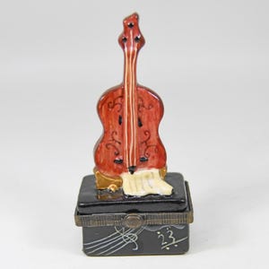 May include: A miniature ceramic figurine of a cello in shades of red and brown, atop a black box with musical notes and the number 23. The cello has white accents and is set on a gold-colored base.