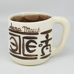 Peut inclure: Mug en céramique crème avec un intérieur marron. Le mug présente des illustrations de style tribal marron et les mots "Hana Maui" en script marron. La poignée est de couleur crème.