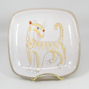 May include: A square ceramic plate with rounded corners featuring a hand-painted yellow tiger illustration. The tiger has thin stripes, a curled tail, and a single blue eye. The plate has a speckled white glaze and a natural tan rim.