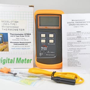 Puede incluir: Termómetro digital naranja y negro con una sonda amarilla, una caja blanca y un manual. El termómetro tiene una pantalla digital y botones. La caja está etiquetada como "Digital Meter". El dispositivo es para medir la temperatura.
