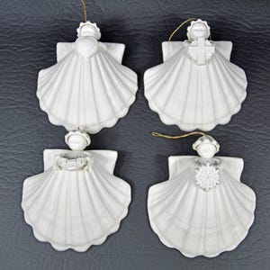 Puede incluir: Cuatro adornos de ángel de cerámica blanca. Cada ángel está hecho de una concha de vieira con una pequeña figura en la parte superior. Los ángeles sostienen un corazón, una cruz, flores y un copo de nieve. Hilo dorado para colgar.