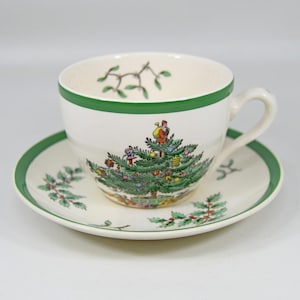 Op de afbeelding: Een witte theekop en schotel set met een groene rand. De theekop heeft een kerstboomontwerp met een kerstmanfiguur. De schotel heeft hulst- en bessenontwerpen. Een feestelijk, vakantie-thema theeservies.