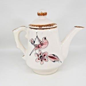 May include: White ceramic teapot with a lid, handle, and spout. The teapot is decorated with a pink bird and flower design. The rim and lid are trimmed with a brown color.