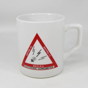 以下が含まれることがあります： 赤い警告三角形が付いた白いセラミックマグ。三角形には稲妻のグラフィックと「AN/FRD-10」、「AN/FRD-13」、「AN/FLR-9」、「ELECTRONIC GONIOMETER」の文字が含まれています。マグには湾曲したハンドルが付いています。