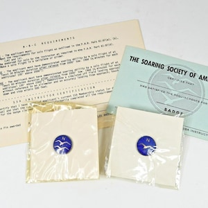 May include: An assortment of documents and badges related to the Soaring Society of America. Includes a light blue certificate, a beige document with text, and two badges with a blue and white emblem.