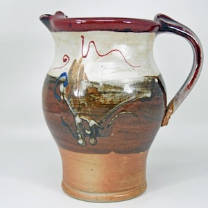 Könnte beinhalten: Handgefertigter Keramikkrug mit breitem Boden und gebogenem Henkel. Der Krug hat eine Farbpalette aus Braun, Creme und Burgunderrot mit abstrakten Malereidetails. Der Krug ist ein dekorativer Artikel für Küche oder Esszimmer.
