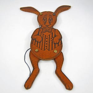 Puede incluir: Un juguete de cuerda de madera vintage con forma de conejo. El conejo marrón tiene un chaleco a rayas negras y marrones y una pajarita. El juguete tiene remaches de latón y una cuerda negra.