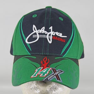 May include: A navy blue and green baseball cap featuring the white embroidered text John Force Racing on the front panel. The brim displays a red flame graphic above the number 14 and a silver X-shaped logo with the text John Force.